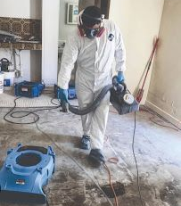Worker in white coveralls uses a floor cleaning machine on a dirty indoor floor.