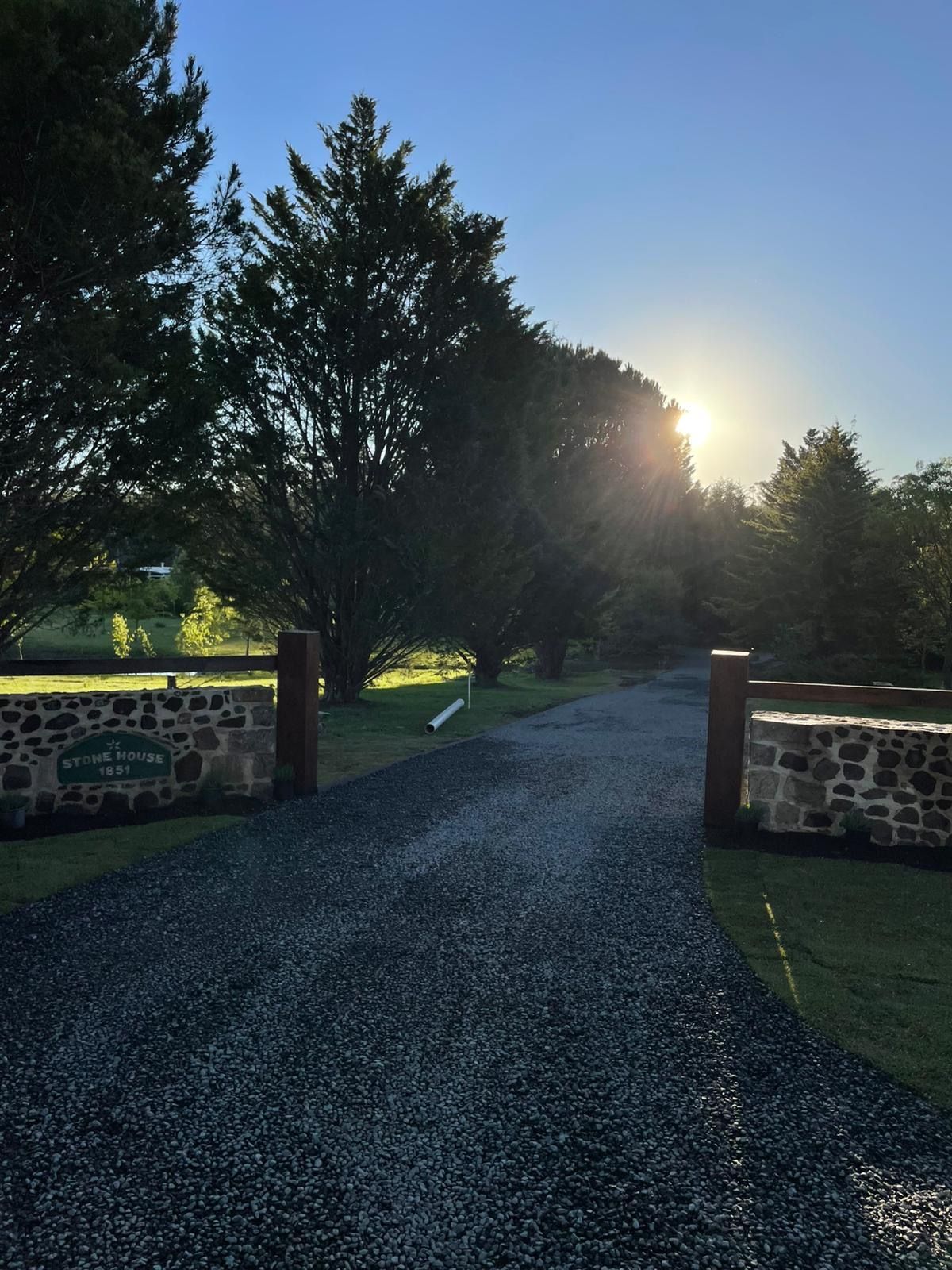 Gravel driveway leads toward sun through trees. Stone pillars frame the entrance.