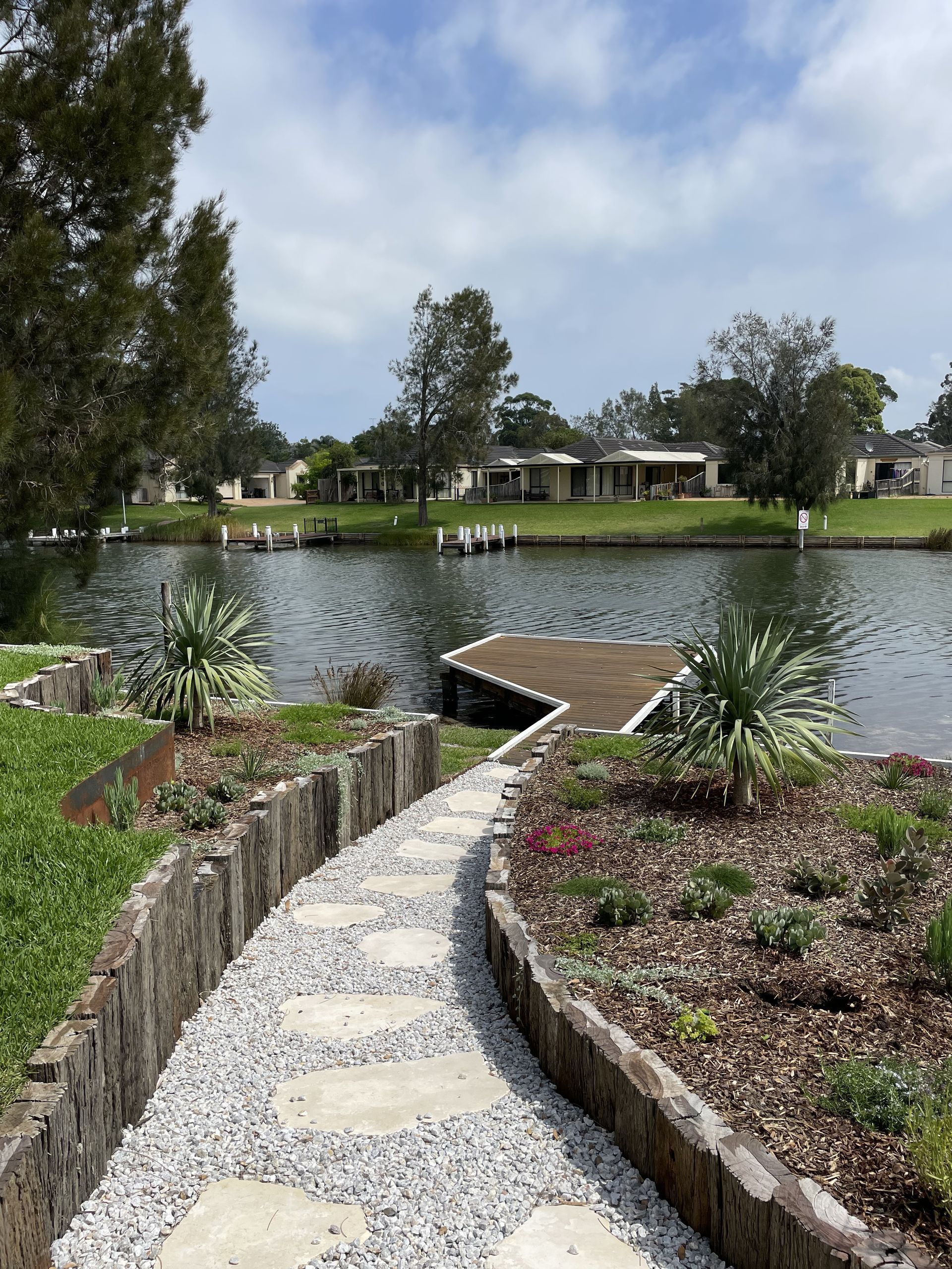 Stone path alongside a lake with a wooden dock and houses in the background under a partly cloudy sky — Schutz Landscape and Garden Supplies in Tomerong, NSW