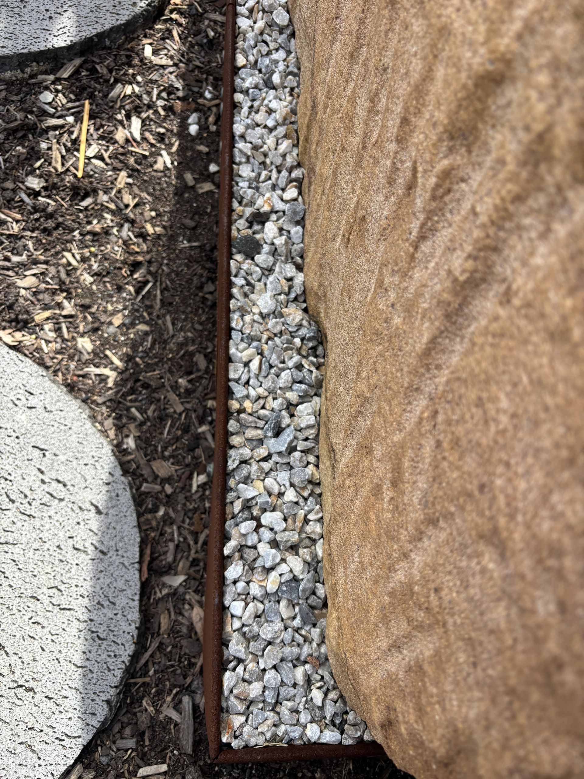 Gravel bed bordered by brown metal, next to a large brown rock and a gray stepping stone, with mulch on left   — Schutz Landscape and Garden Supplies in Tomerong, NSW