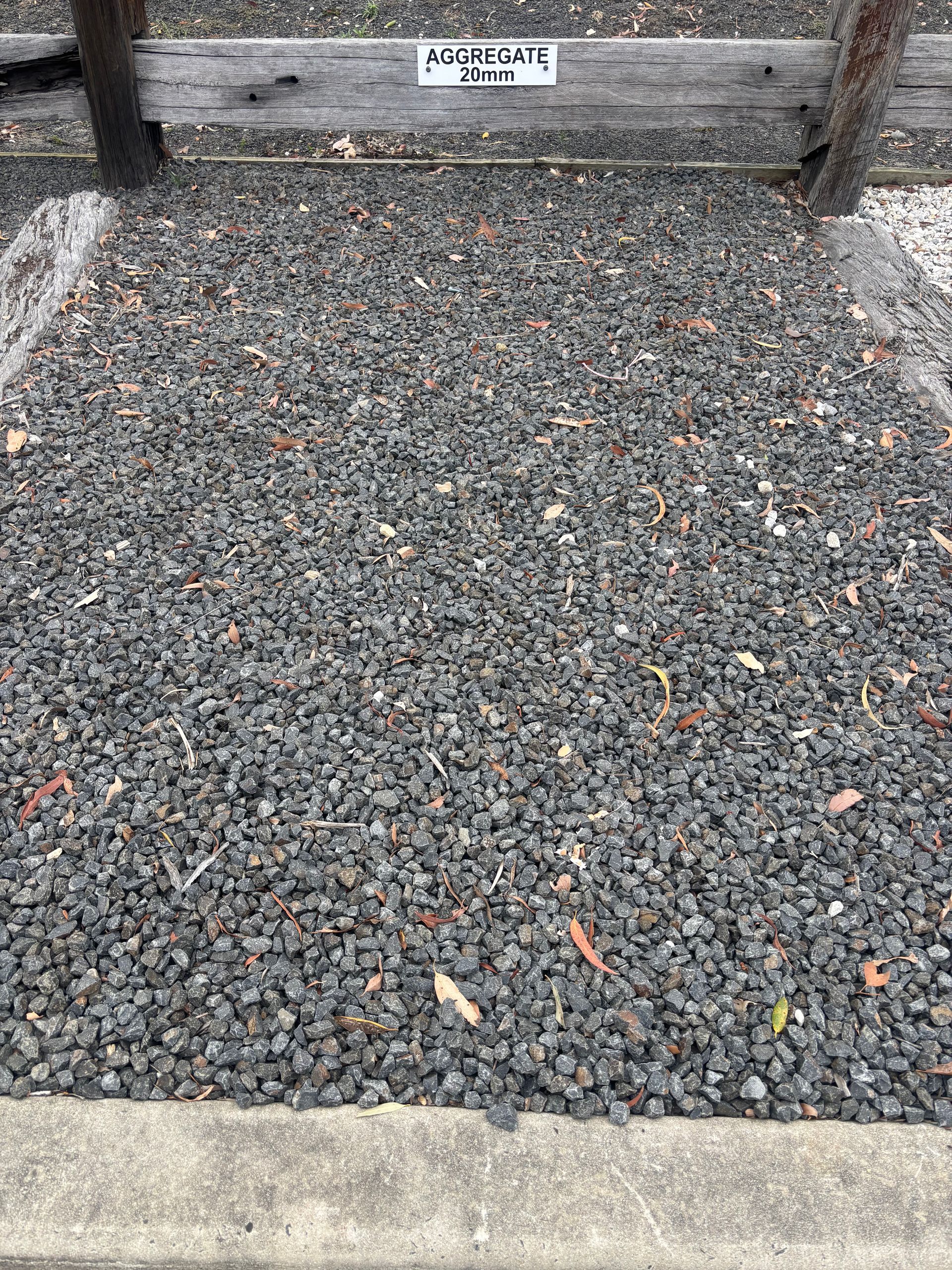 Gravel-covered patch bounded by wooden beams and a concrete edge. A sign is visible on the beams — Schutz Landscape and Garden Supplies in Tomerong, NSW