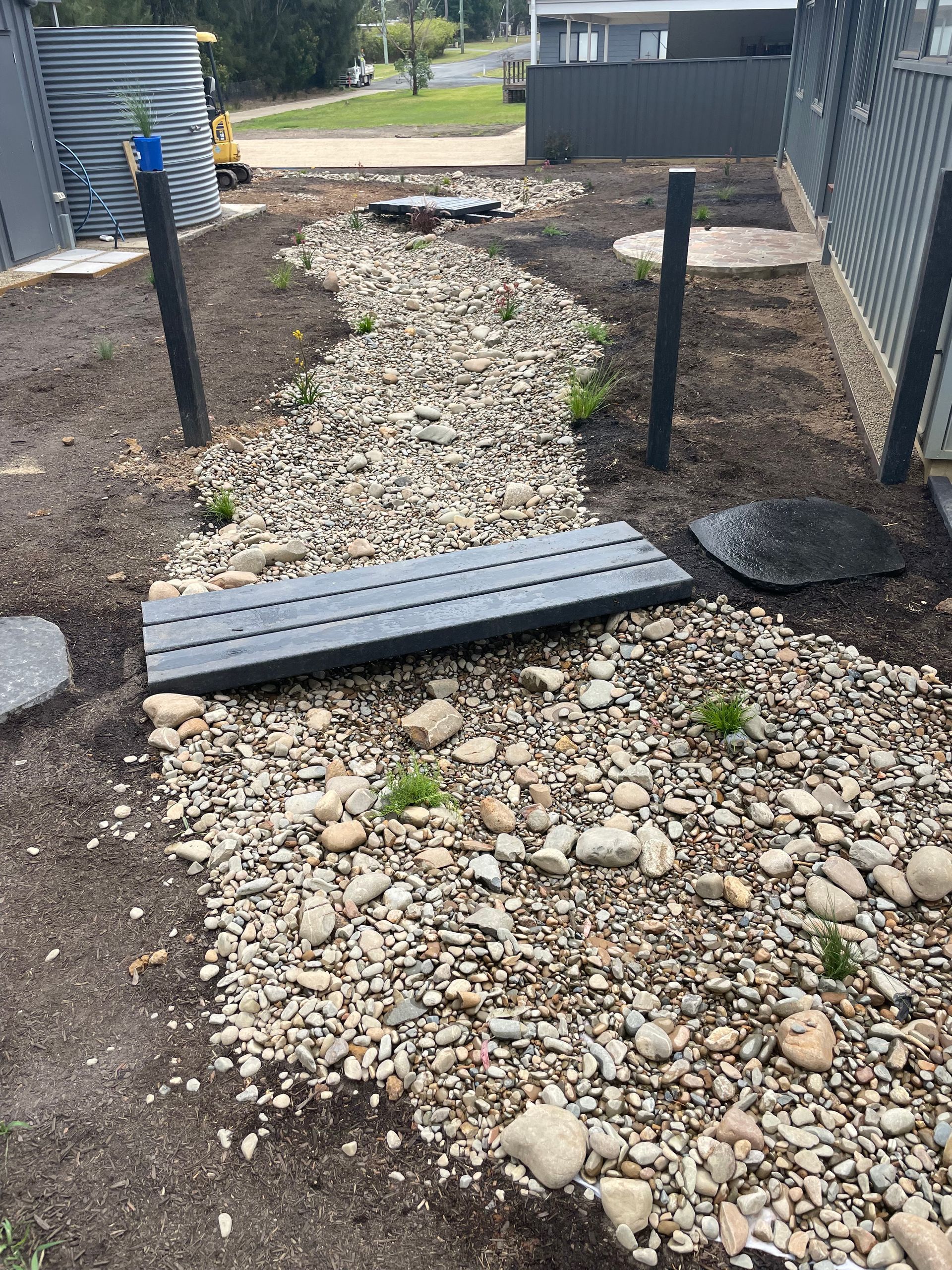 Gravel pathway with small bridge and black posts in a garden setting, leading to a house.