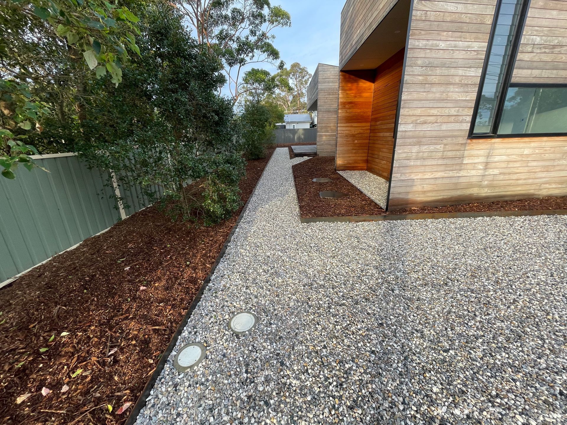 Gravel pathway beside a modern brick building. Mulch bed next to a green fence, trees in the background.