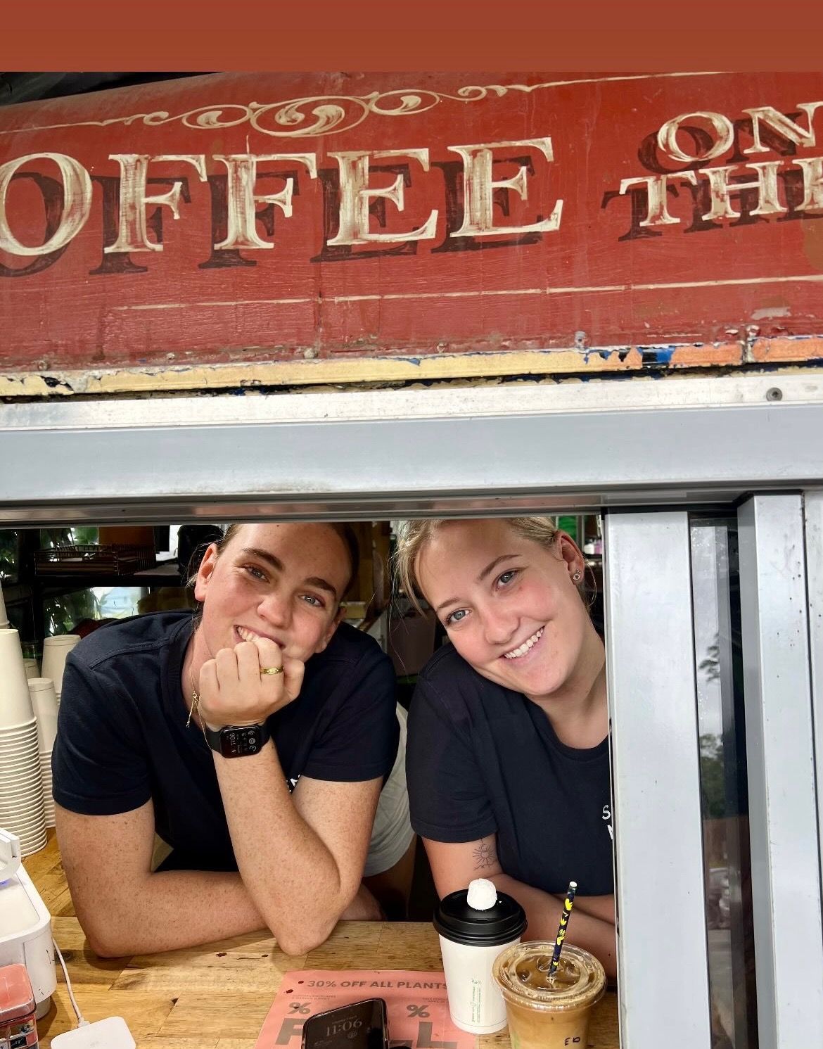 Two people smile from a coffee stand window. A sign reads