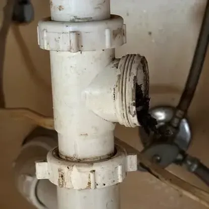 A close-up of a white plastic plumbing pipe under a sink with a detached, open joint revealing dark debris inside.