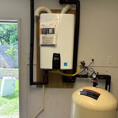 A white tankless water heater mounted on a plywood board next to a beige expansion tank in a utility room.