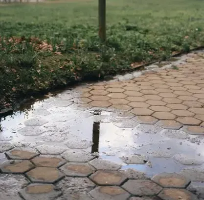 A puddle of water sits on a hexagonal paved path next to a grassy lawn, reflecting a nearby post.