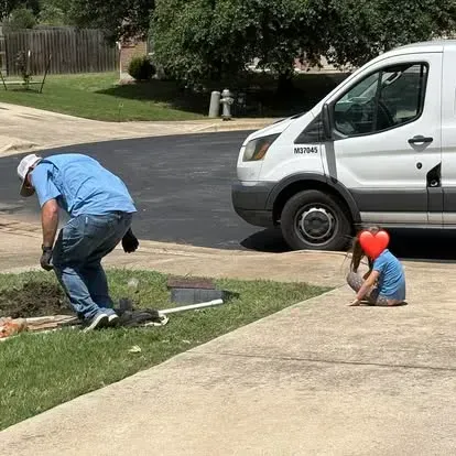 A worker in a blue shirt and jeans digs in a lawn near a white van, while a child sits on a driveway nearby.