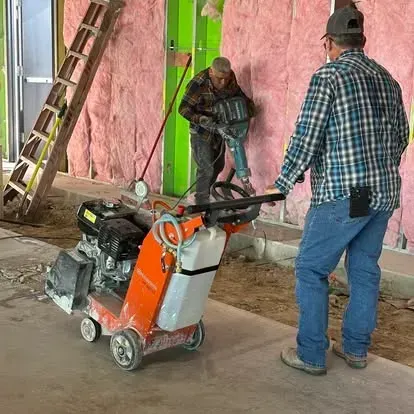 Two workers operate construction equipment inside a building with exposed pink insulation walls and a wooden ladder.