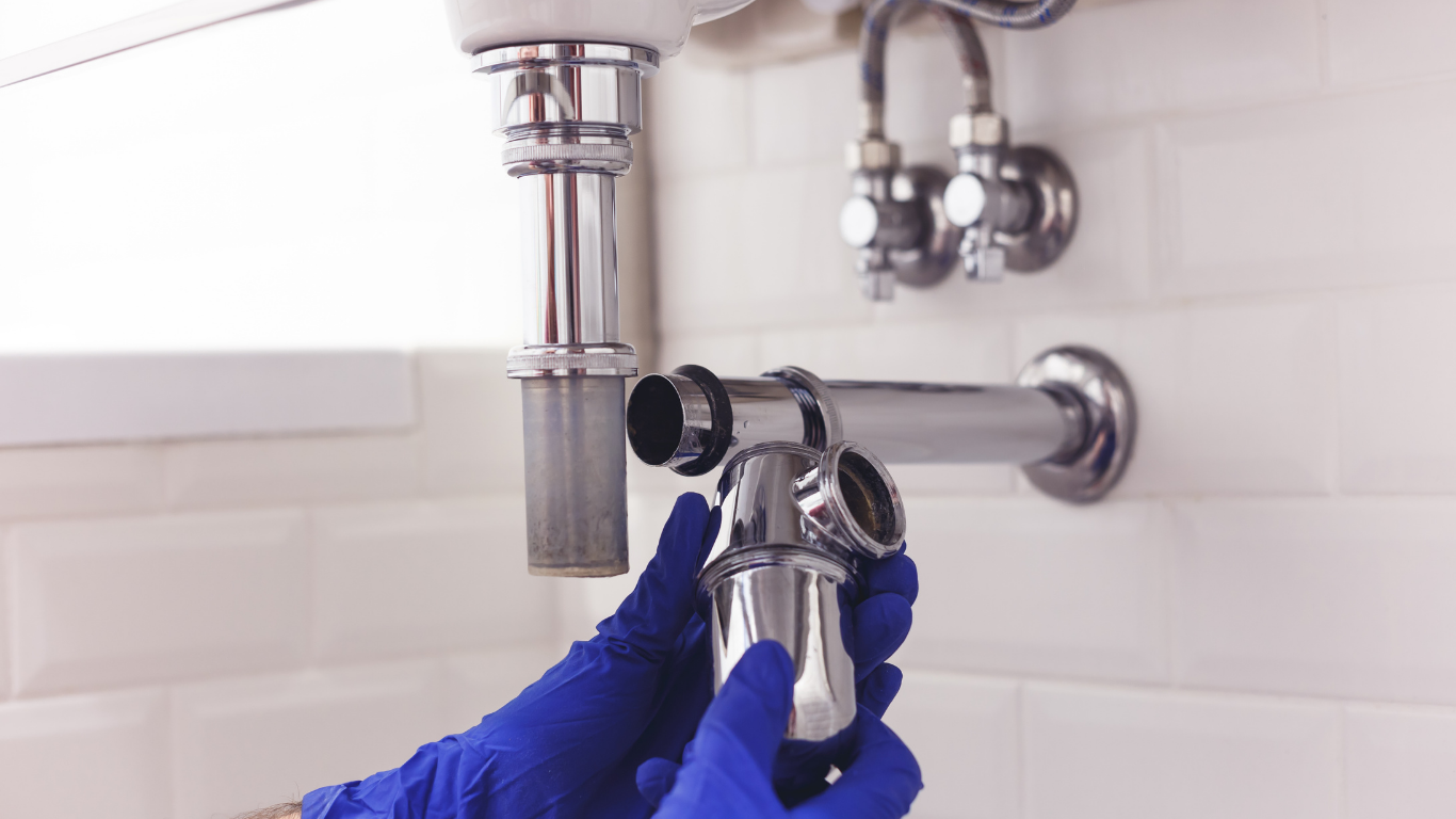 A person wearing blue gloves works to install a chrome p-trap under a bathroom sink against a white tiled wall.