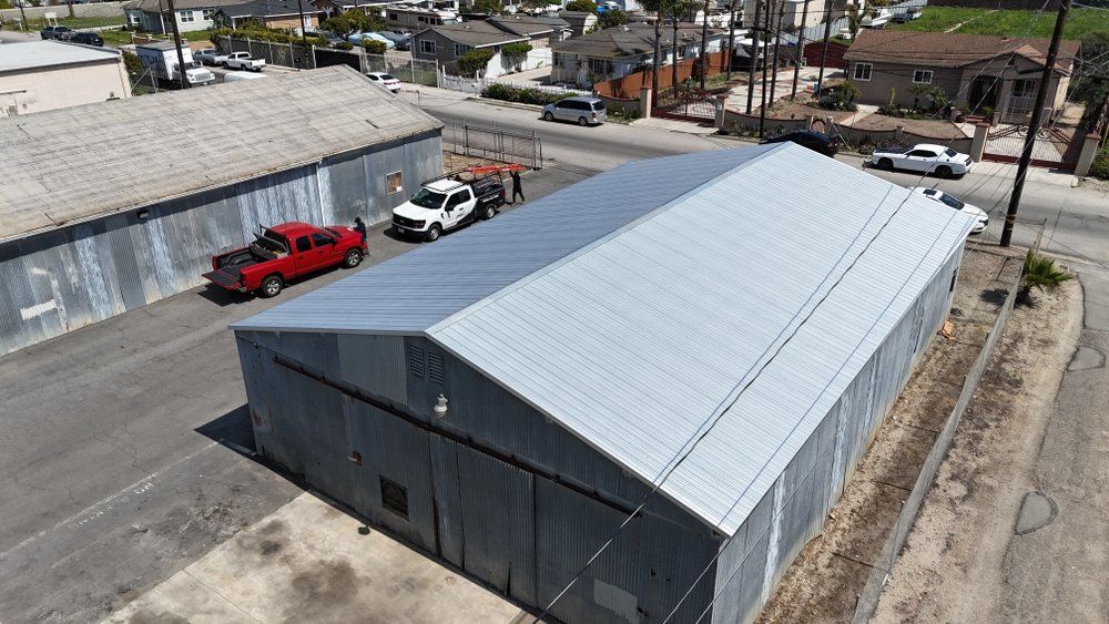 Close-up of a dark grey metal roof with a ridged design. Below the roof is a white trim and brick exterior. Close-up of a dark grey metal roof with a ridged design. Below the roof is a white trim and brick exterior.