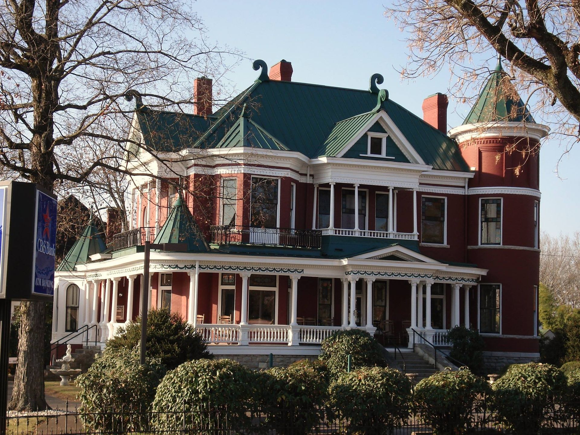 A grand Victorian-style house with red brick walls, a green roof, a turret, and a wrap-around porch.