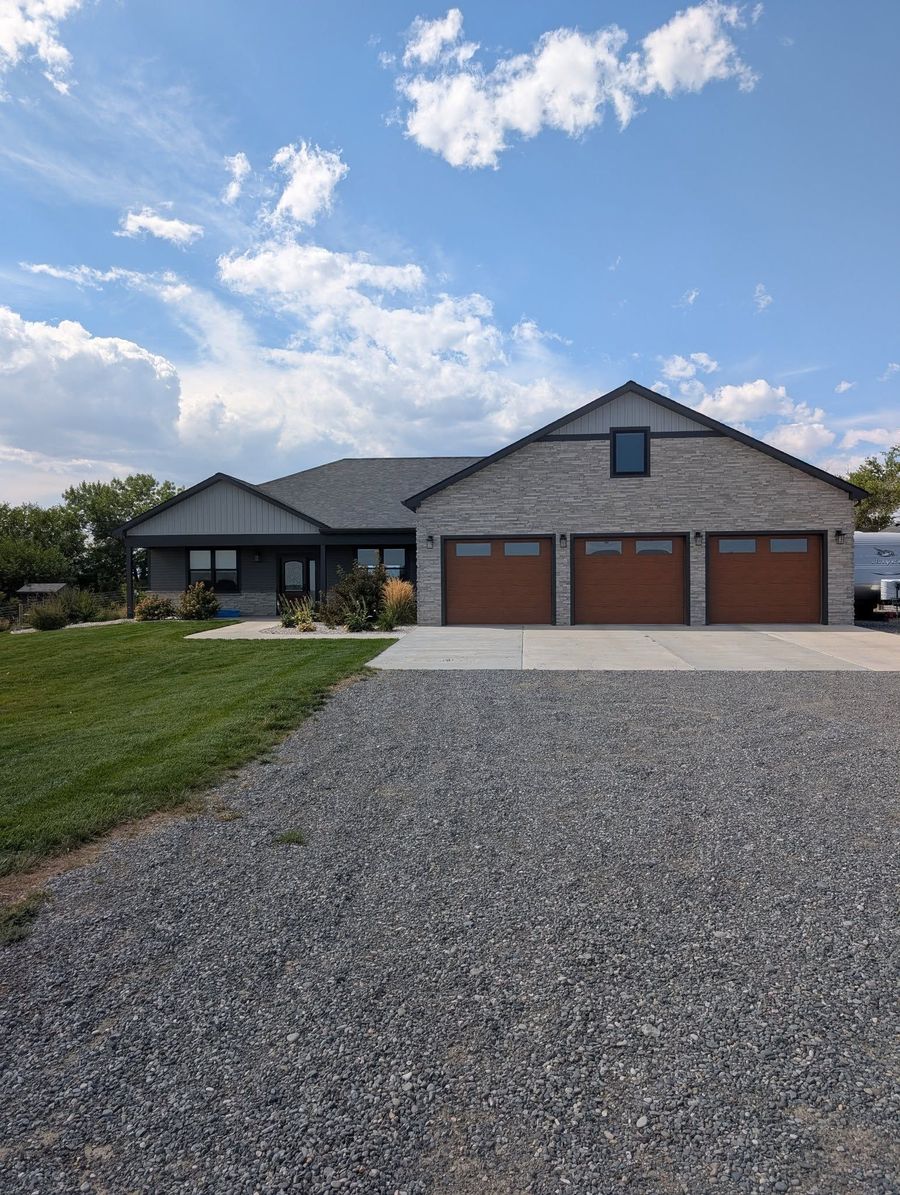 A modern, stone-clad house with a three-car garage and gravel driveway under a blue, cloudy sky.