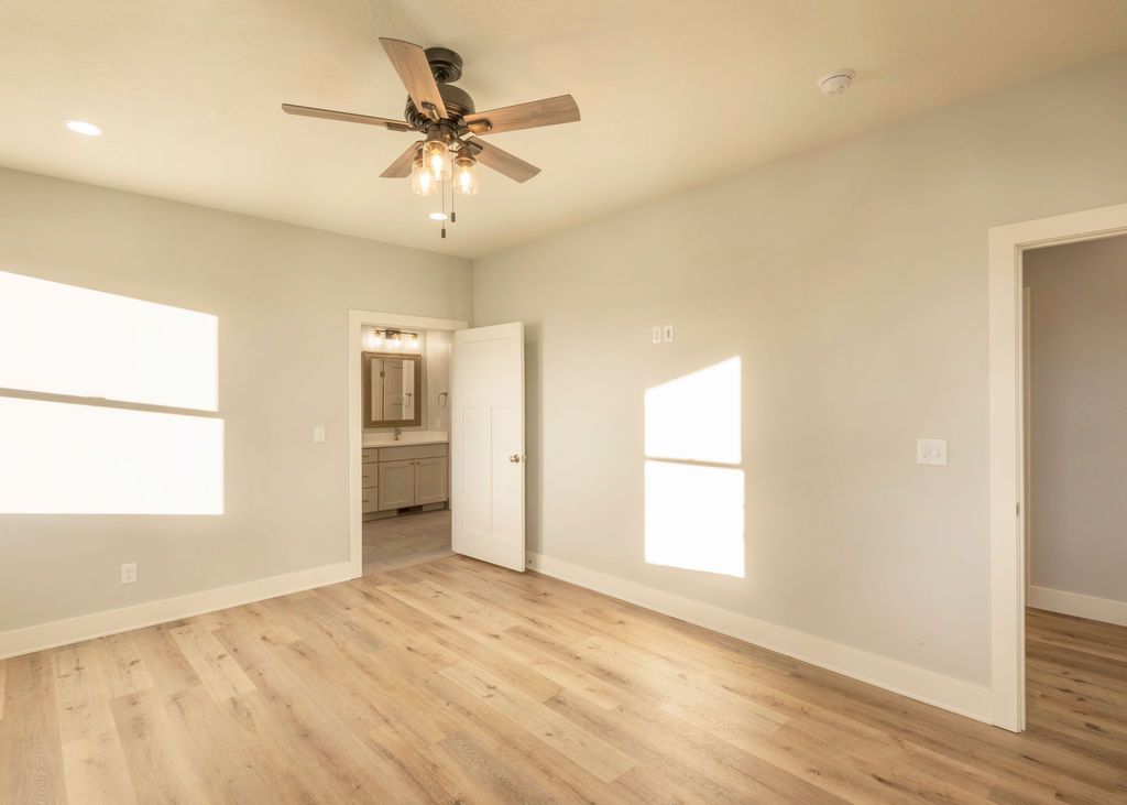 Empty bedroom with light wood floor, two windows, open doorway to a bathroom, and ceiling fan.