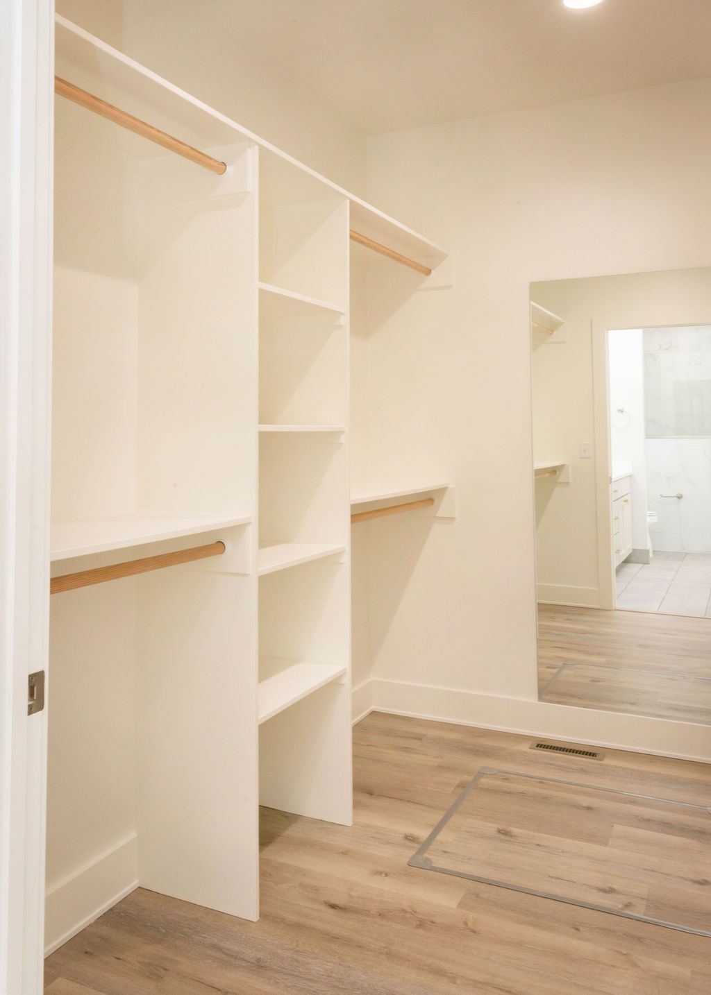 Empty white closet with shelves and hanging rods; wood-look flooring; door leads to bedroom.