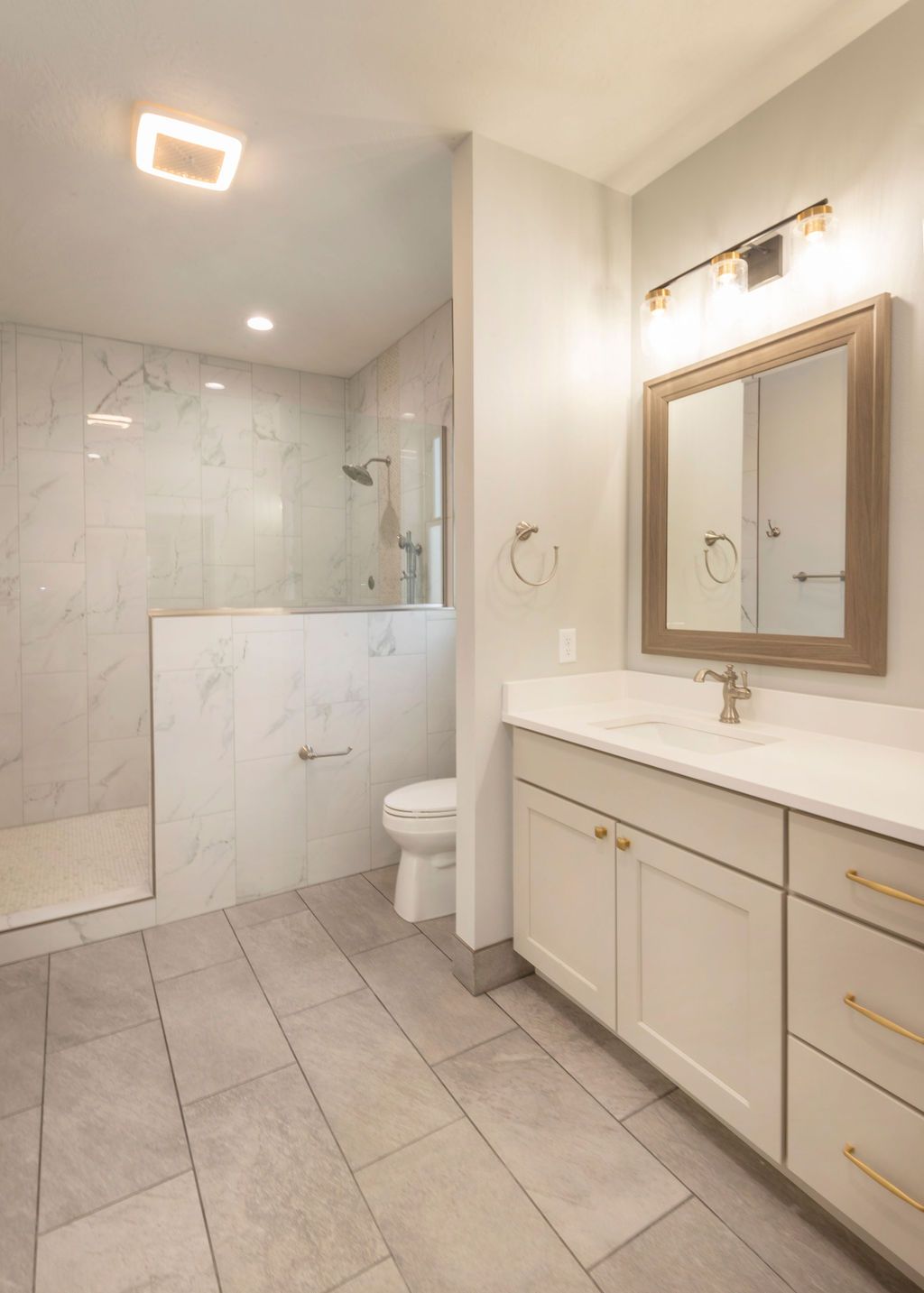 Bright, modern bathroom with walk-in shower, white vanity, toilet, and large mirror. Light gray tiled floor and walls.