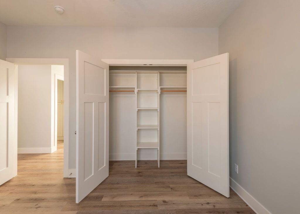 Empty closet with open white doors and shelves, in a room with wood floors and neutral walls.