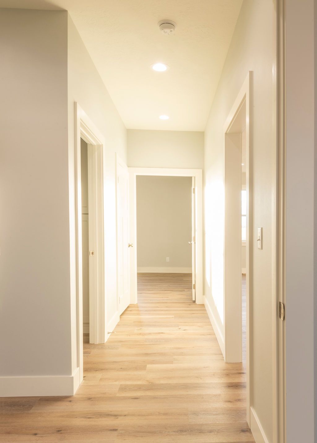 A long hallway with light wood floors and doors leading to rooms, all painted white.
