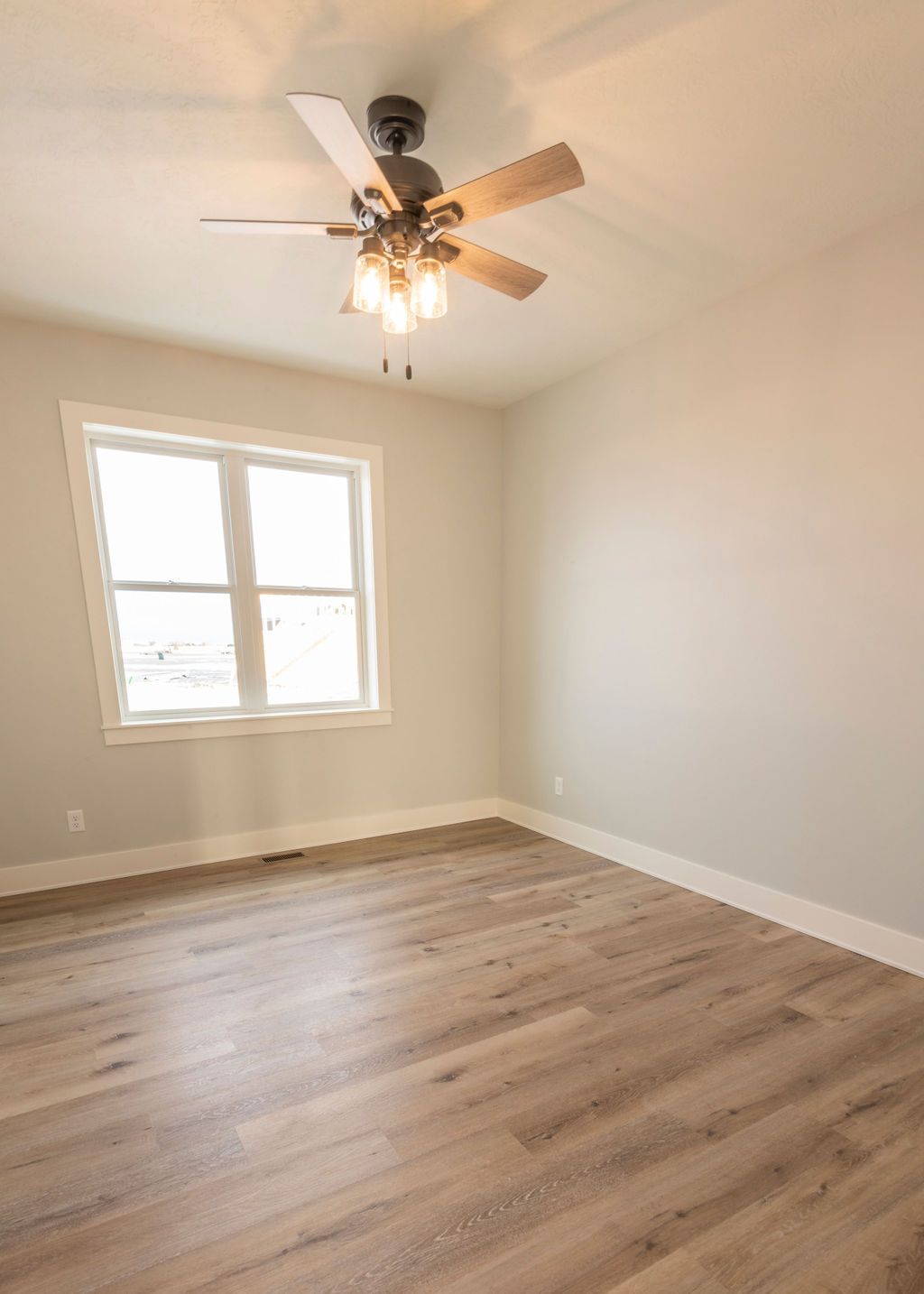 Empty bedroom with wood floor, window, ceiling fan, and neutral-colored walls.