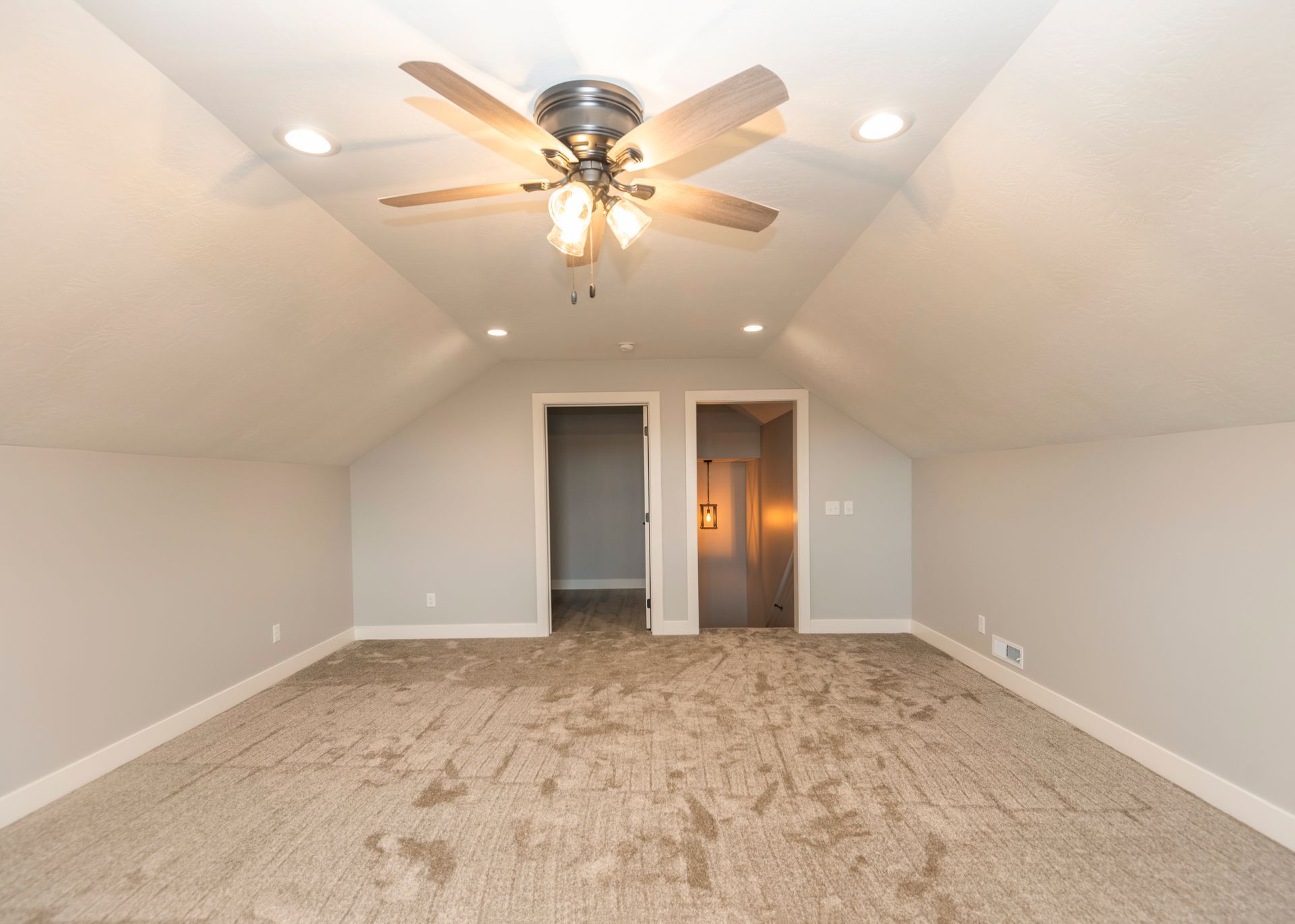 Empty room with sloped ceiling, recessed lights, ceiling fan, and two doorways. Carpeted floor, neutral colors.