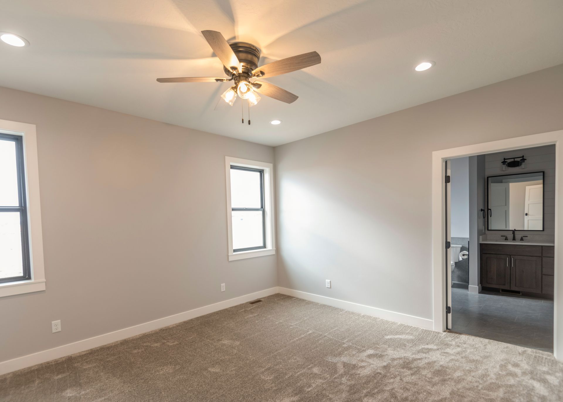 Empty bedroom with beige carpet, light gray walls, and a ceiling fan; doorway to a bathroom.
