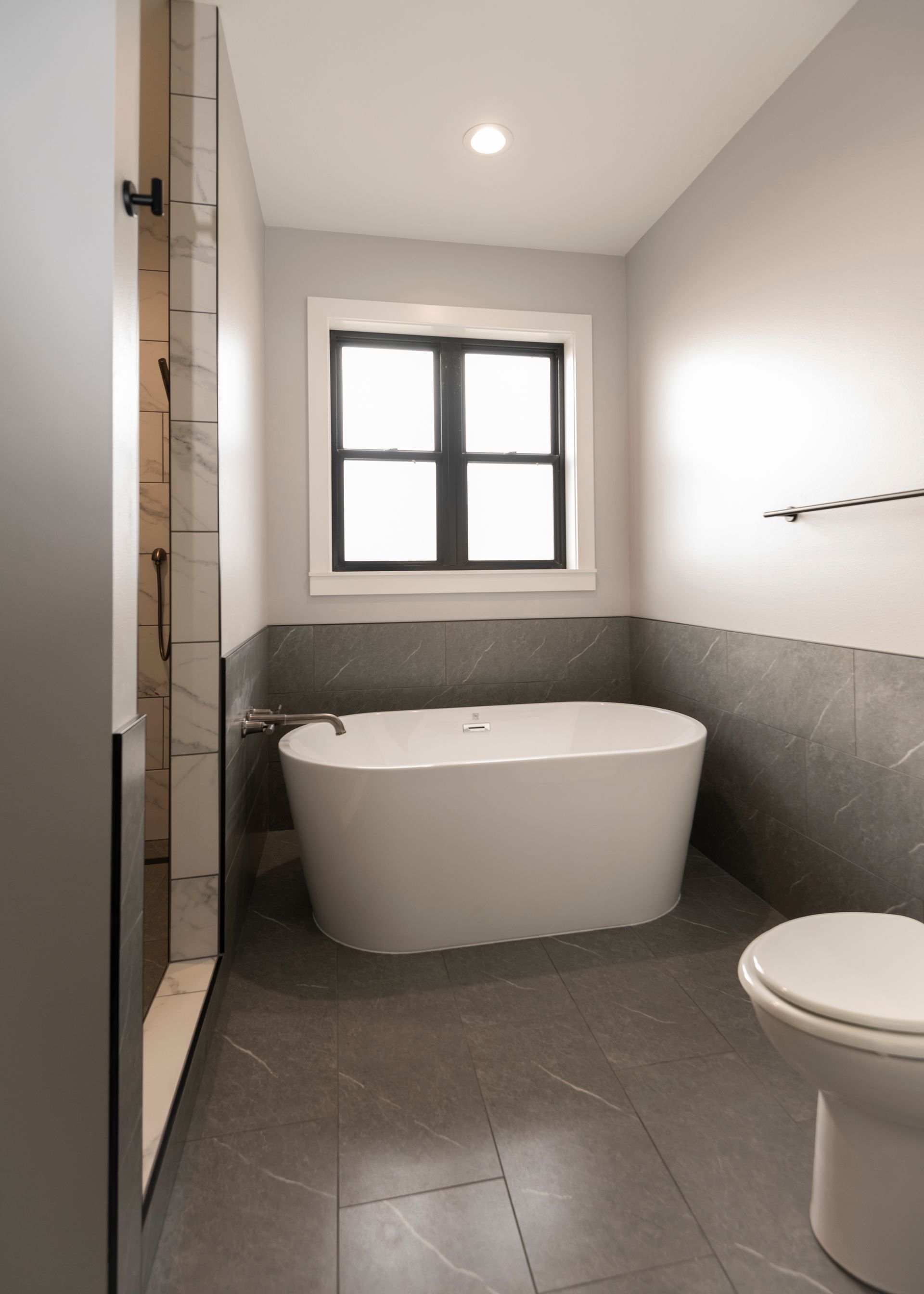 Modern bathroom with white tub, toilet, gray tile, shower, and black-framed window.