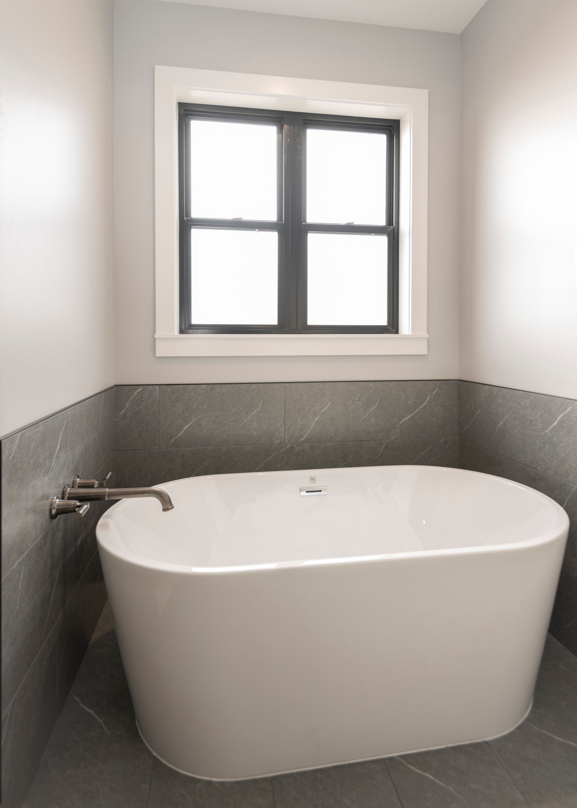 Freestanding white bathtub in a modern bathroom with dark tile accents and a window.