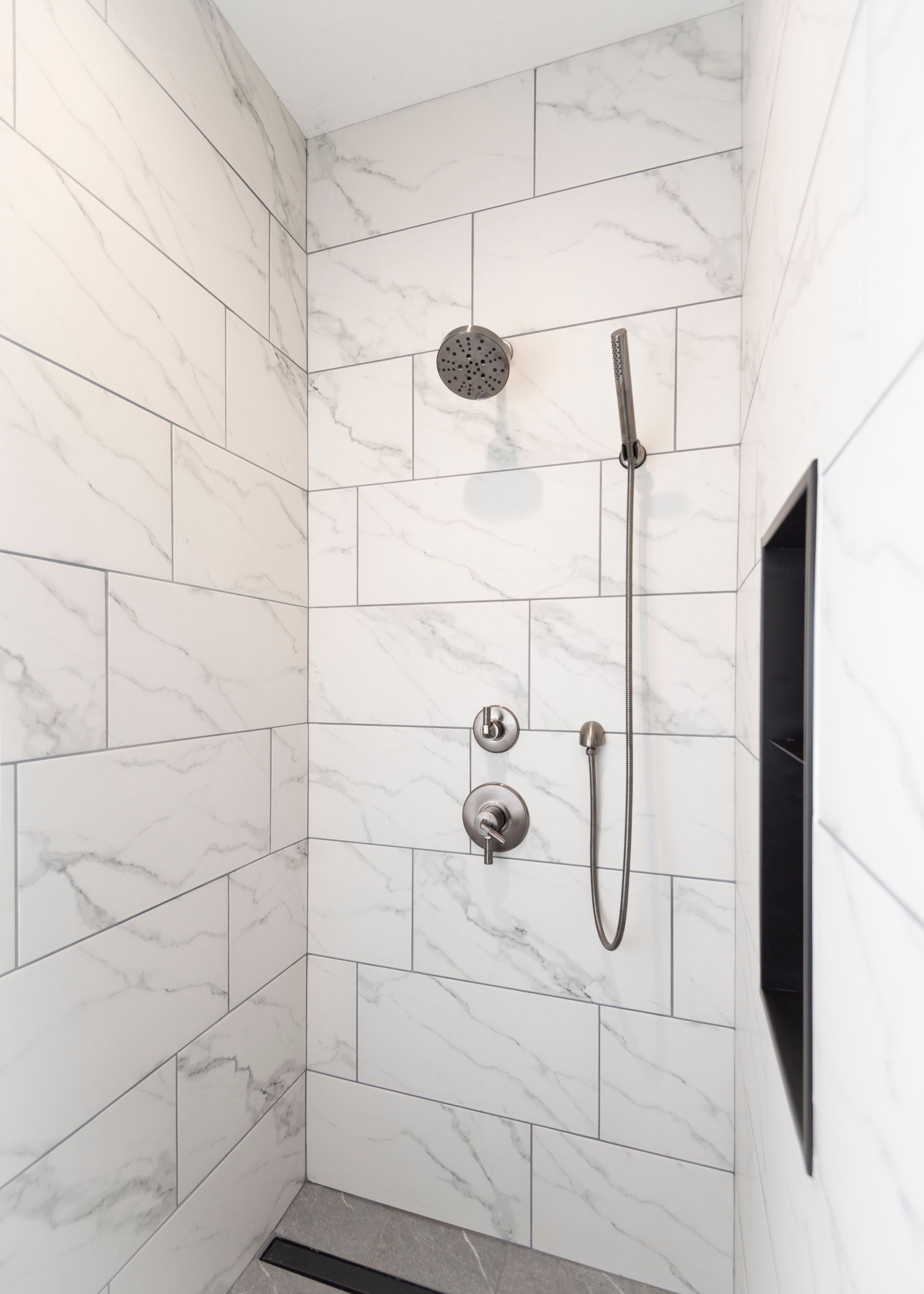 White marble-tiled shower with rain showerhead, handheld wand, control knobs, and a black recessed niche.