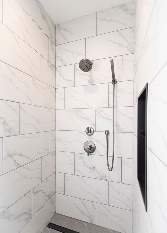 White marble-tiled shower with rain showerhead, handheld wand, control knobs, and a black recessed niche.