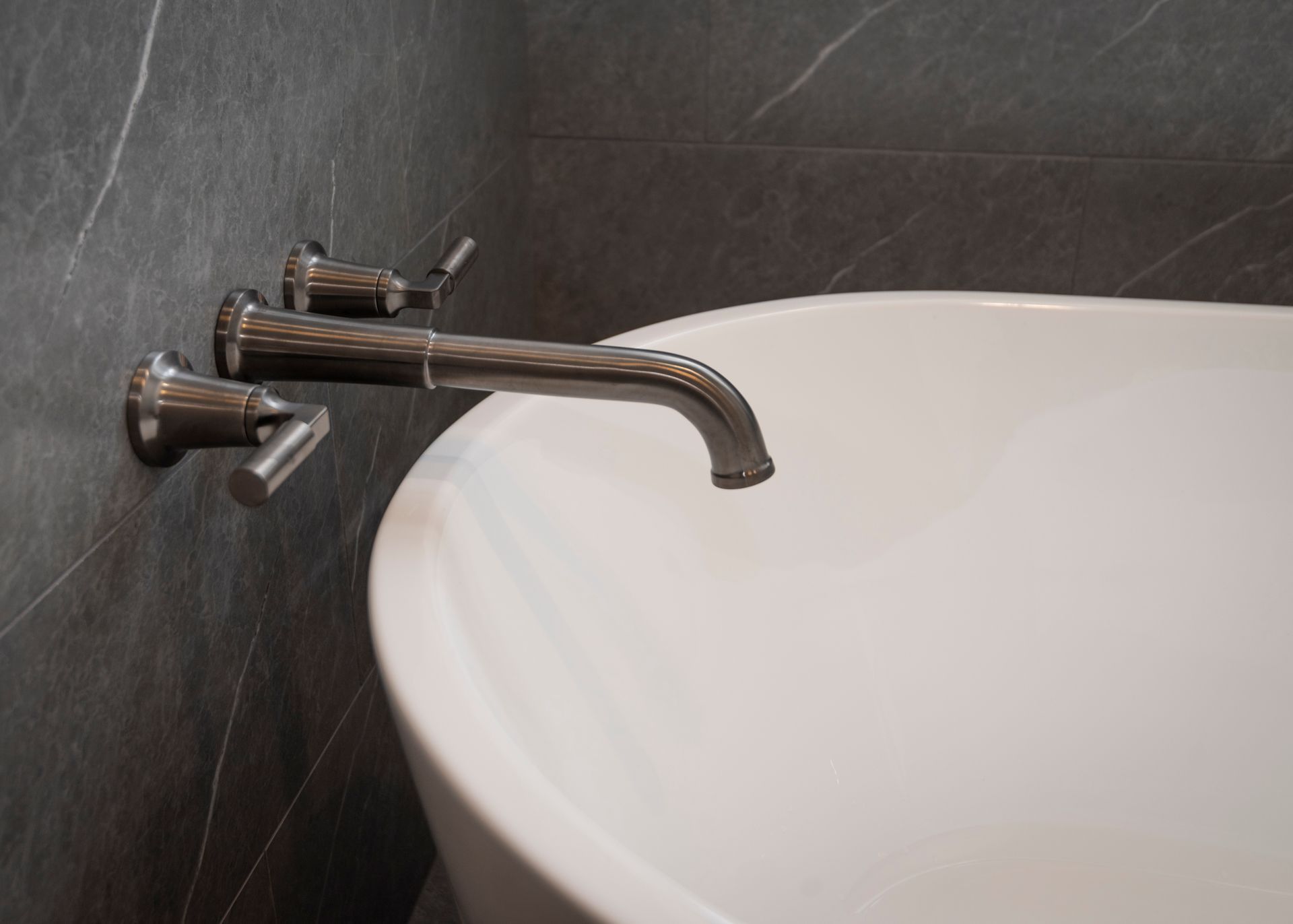 Brushed nickel bathtub faucet over a white tub against a dark gray, marbled wall.