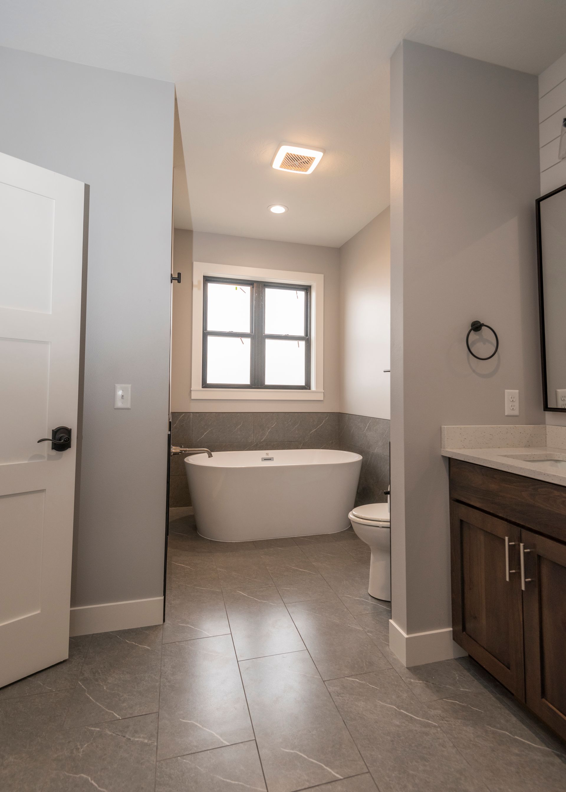 Bathroom with a white tub, toilet, and vanity; grey walls and floor; a window provides light.