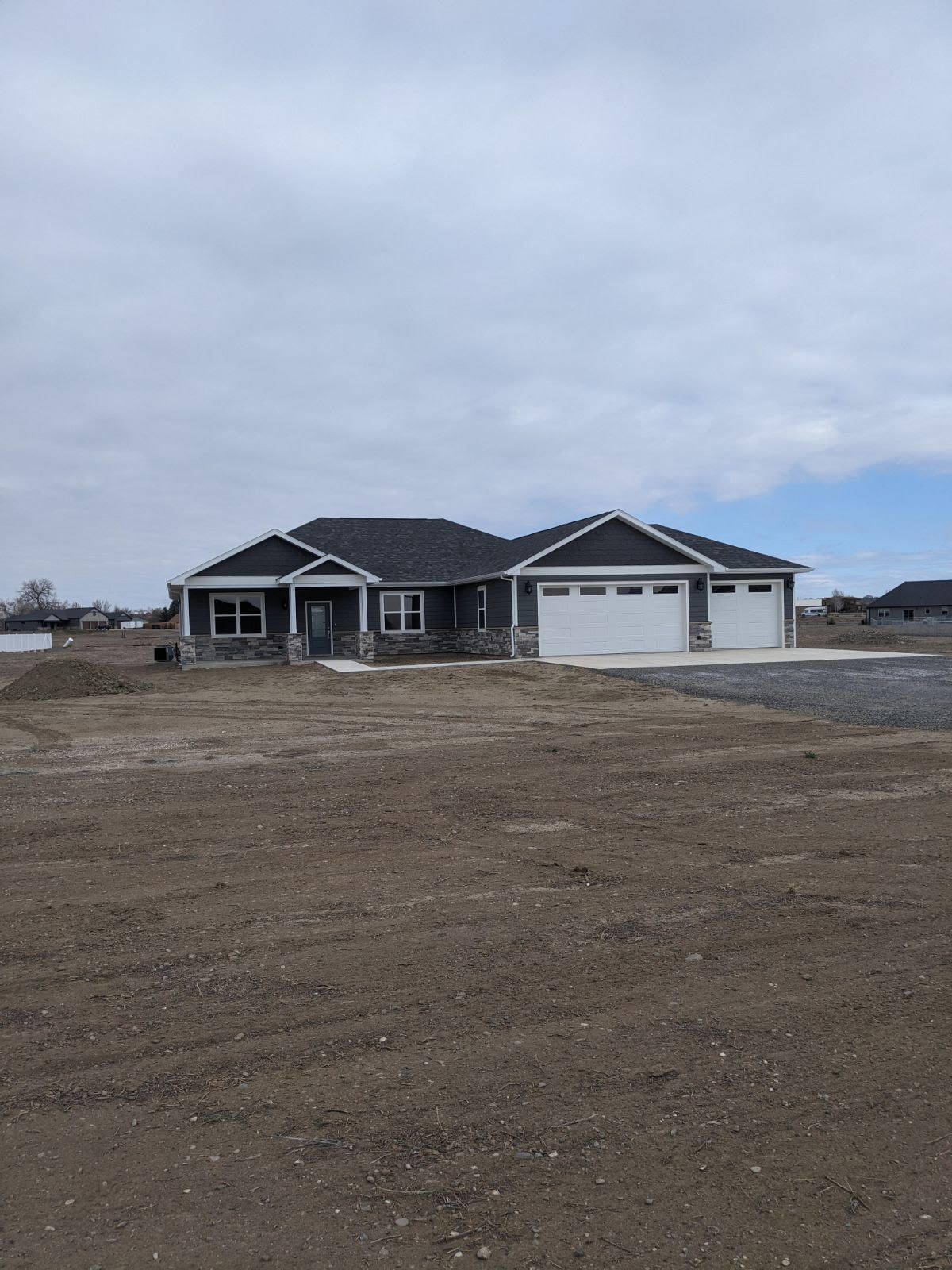 New gray house with white garage doors on a dirt lot under a cloudy sky.