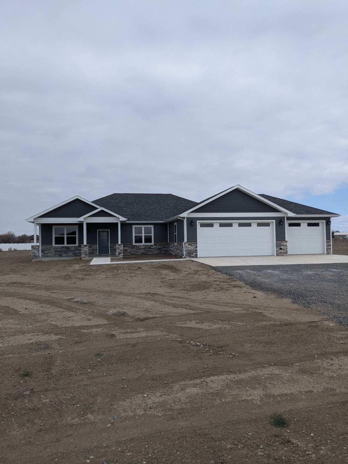 A gray ranch-style house with a two-car garage on a dirt lot under a cloudy sky.