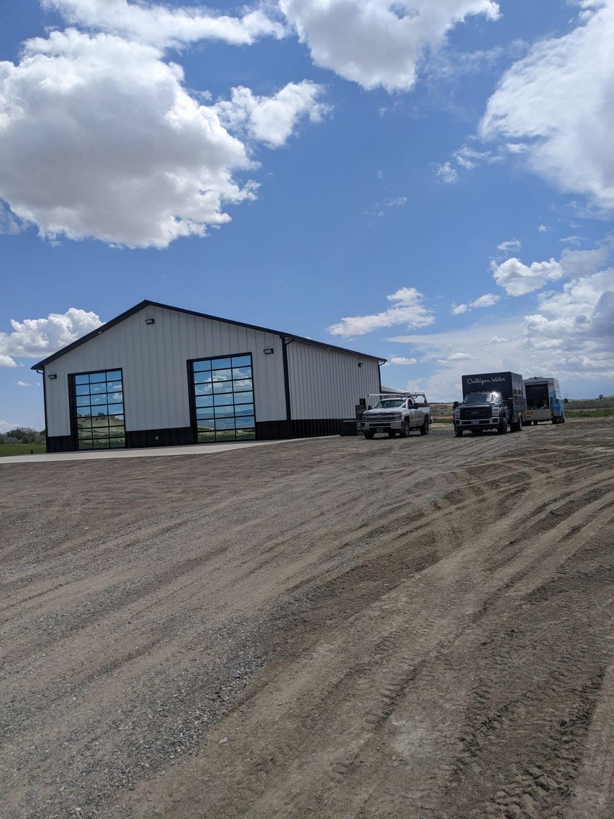 A white metal building with two open doors, vehicles parked outside, under a cloudy blue sky.