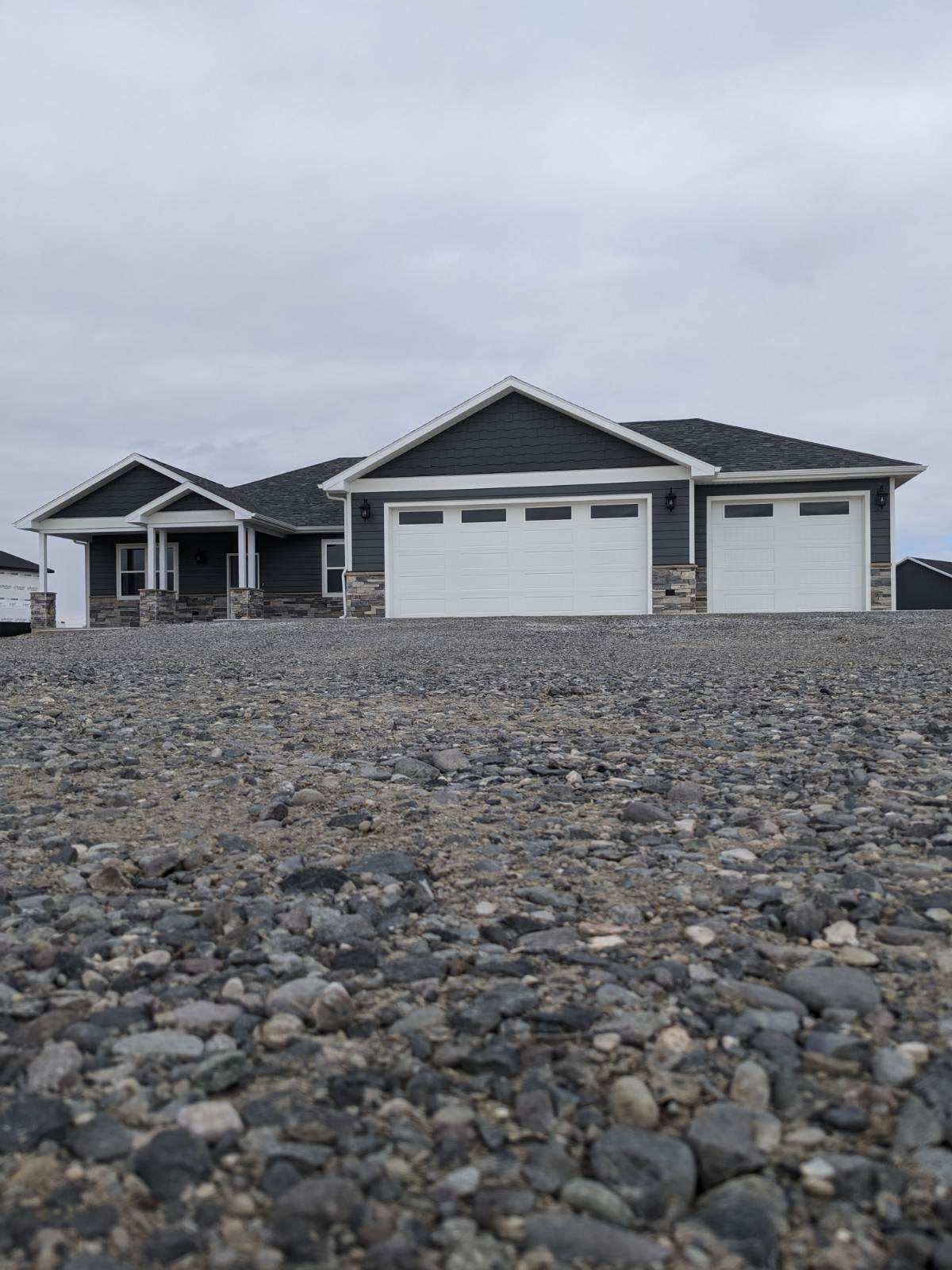 Dark gray house with white garage doors, gray roof, and gravel yard under a cloudy sky.