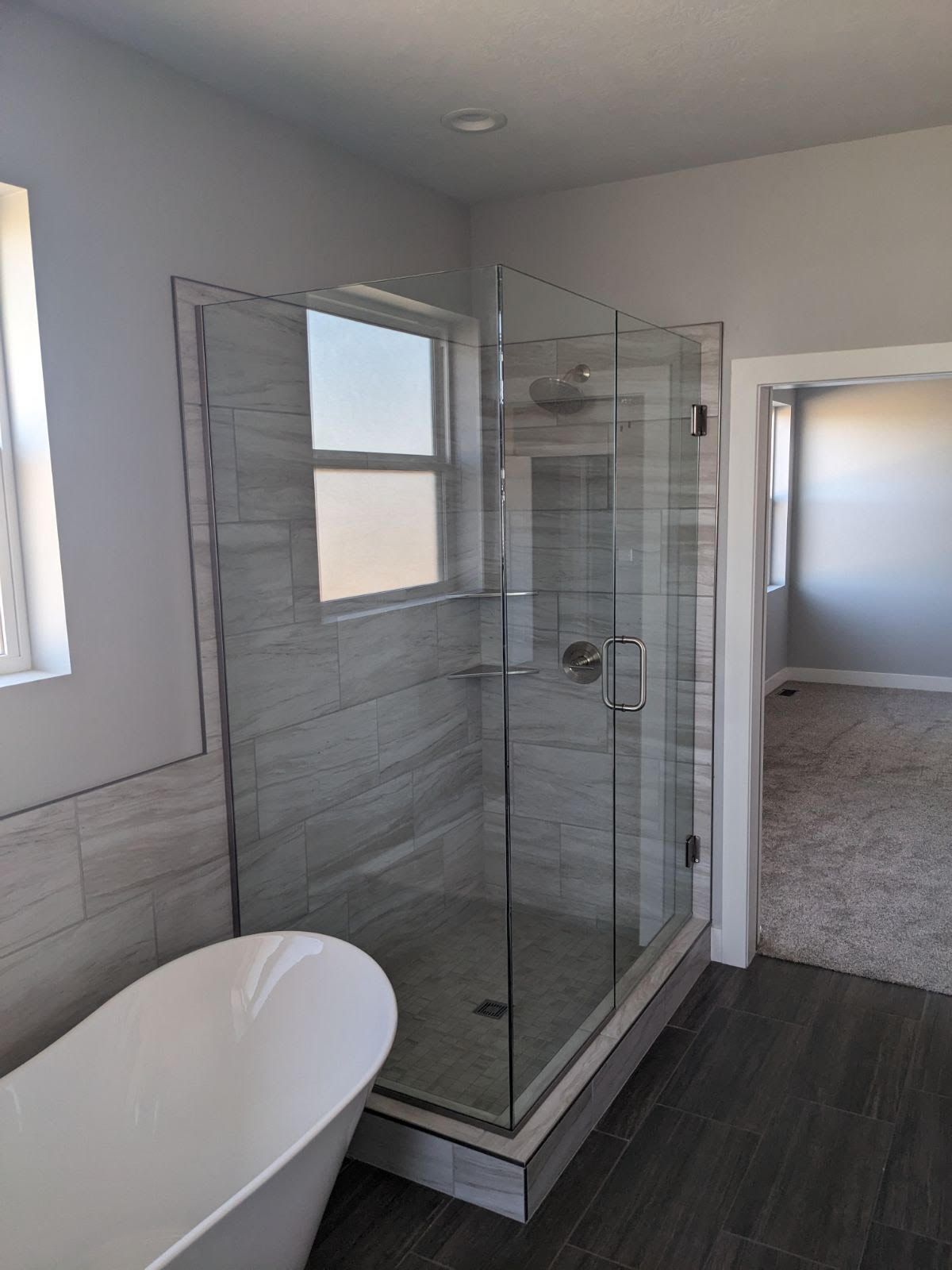 Bathroom with a clear glass shower, bathtub, and doorway to bedroom. Gray and white color scheme.