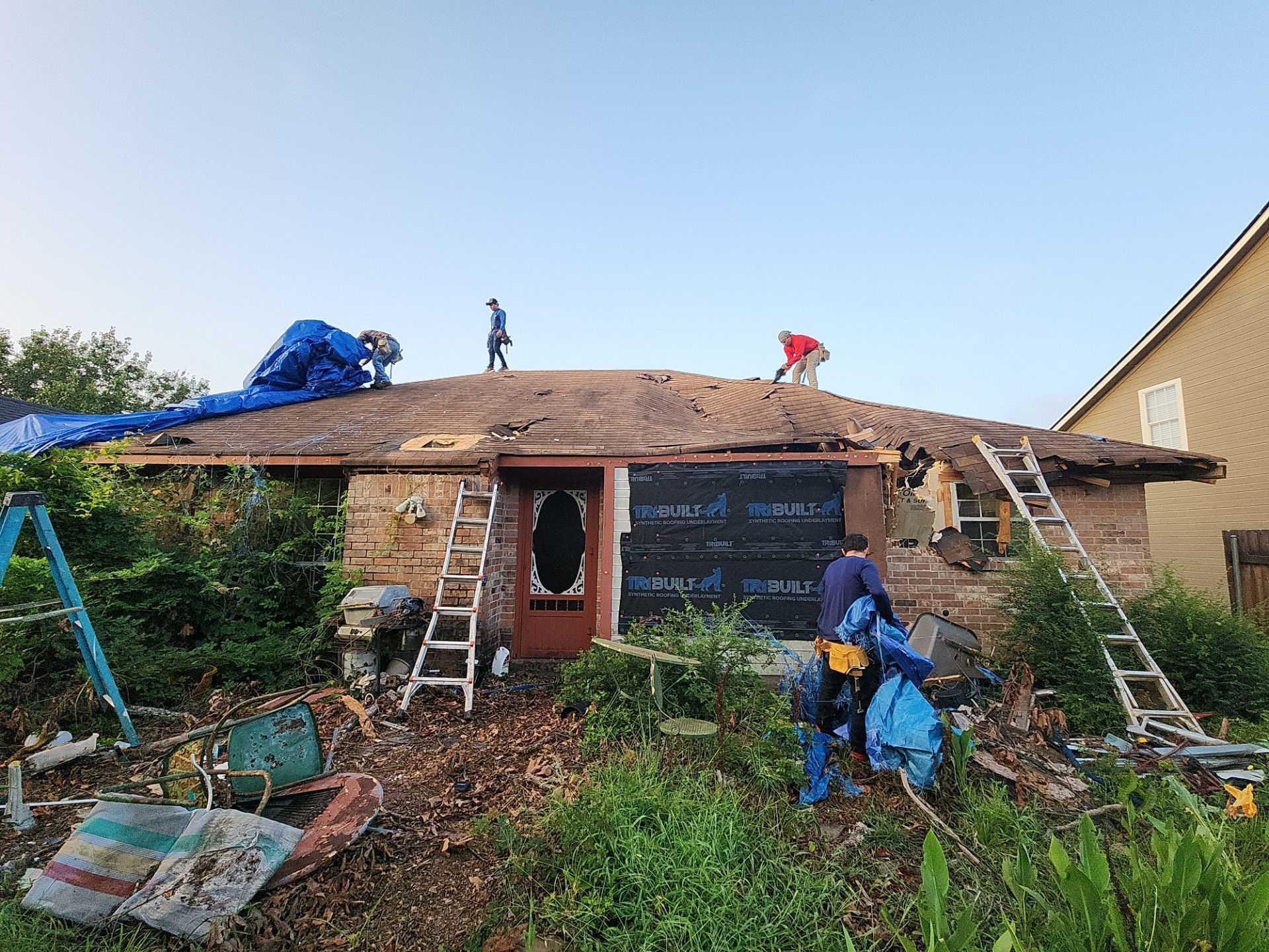 People repairing a damaged house roof. Debris and ladders surround the structure, tarps are placed, and the sky is clear.