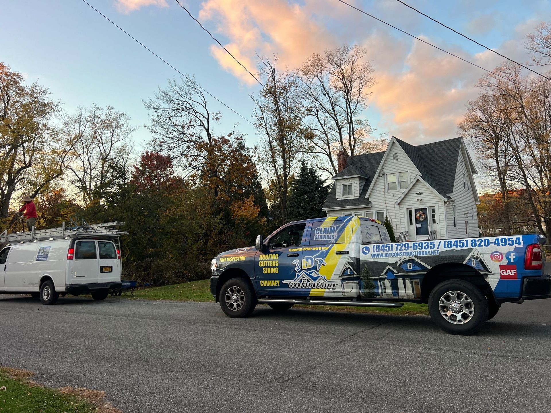 A truck and van parked near a house, possibly a roofing company. Sunset background.