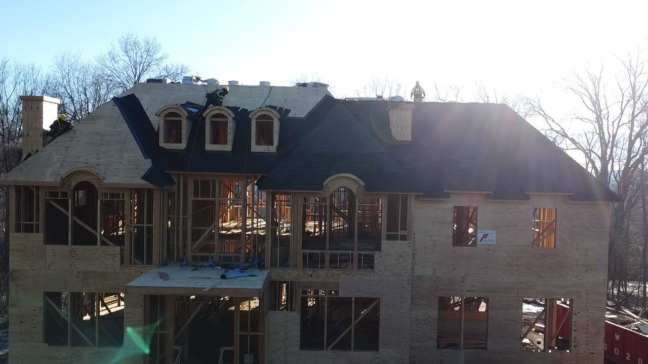 Two-story house under construction with exposed framing and a partially shingled roof. Sunny daytime setting.