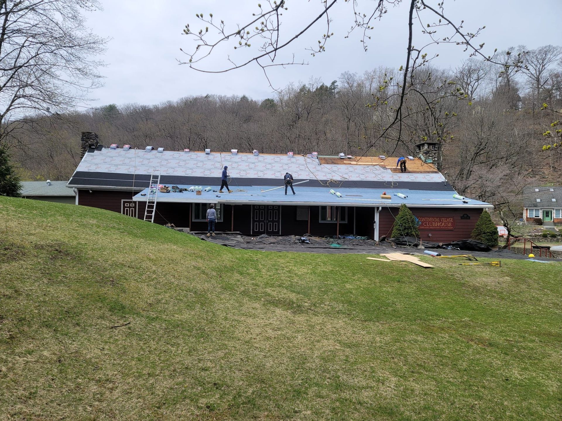 House with roof under construction; workers on roof, blue tarp, green grass, trees.