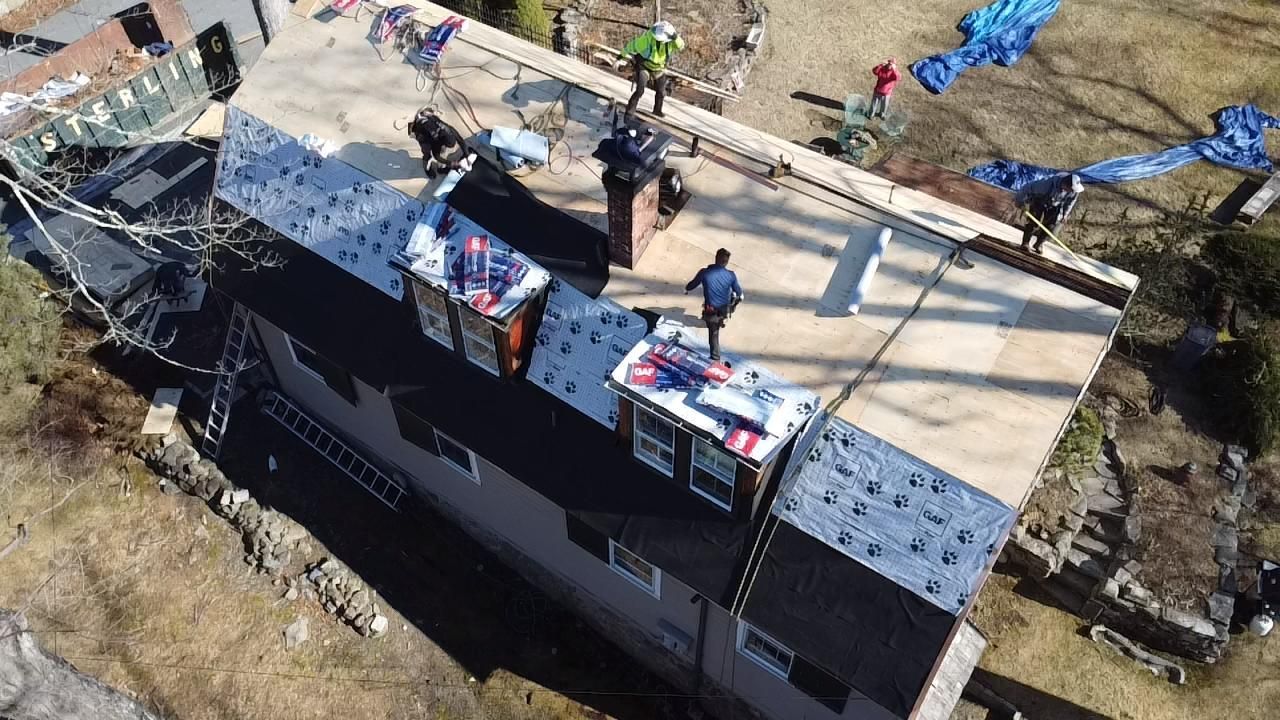 Workers on a roof during a renovation, installing roofing material. Blue tarps cover portions of the roof.
