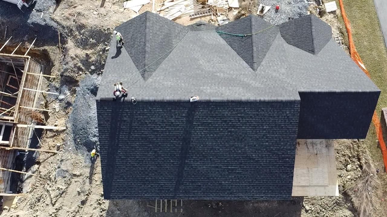 Overhead view of a house under construction with a dark gray shingled roof and workers on top.