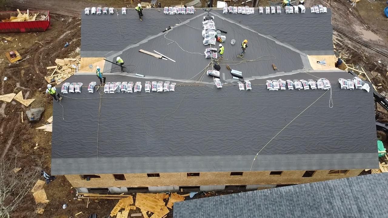Roofers installing shingles on a large, multi-section building. Workers in yellow vests.