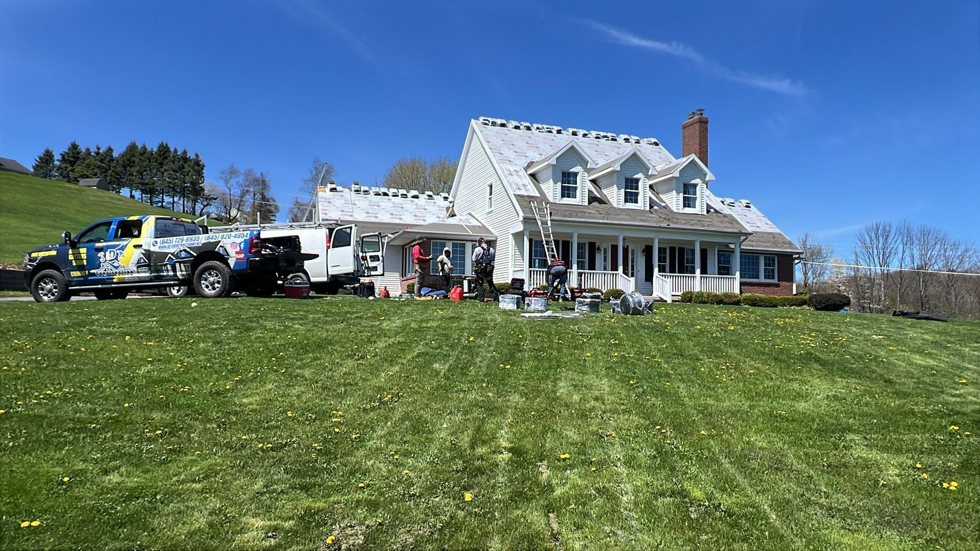 White house with a new roof being installed; construction truck parked on the lawn under a bright blue sky.