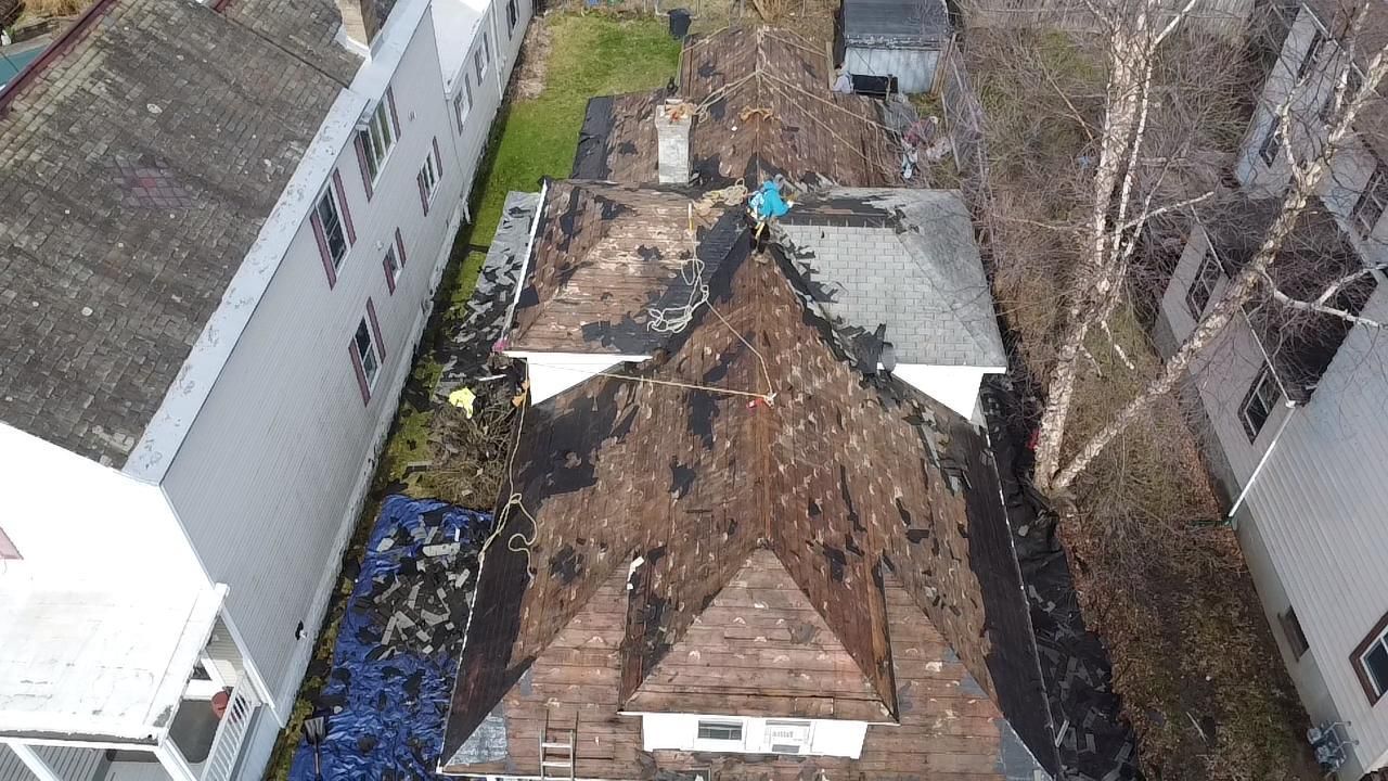 Overhead view of a roof with a person working. Debris covers much of the surface. Buildings flank the home.