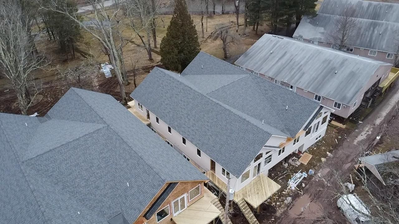 Aerial view of new buildings with gray roofs under construction next to older buildings and a wooded area.