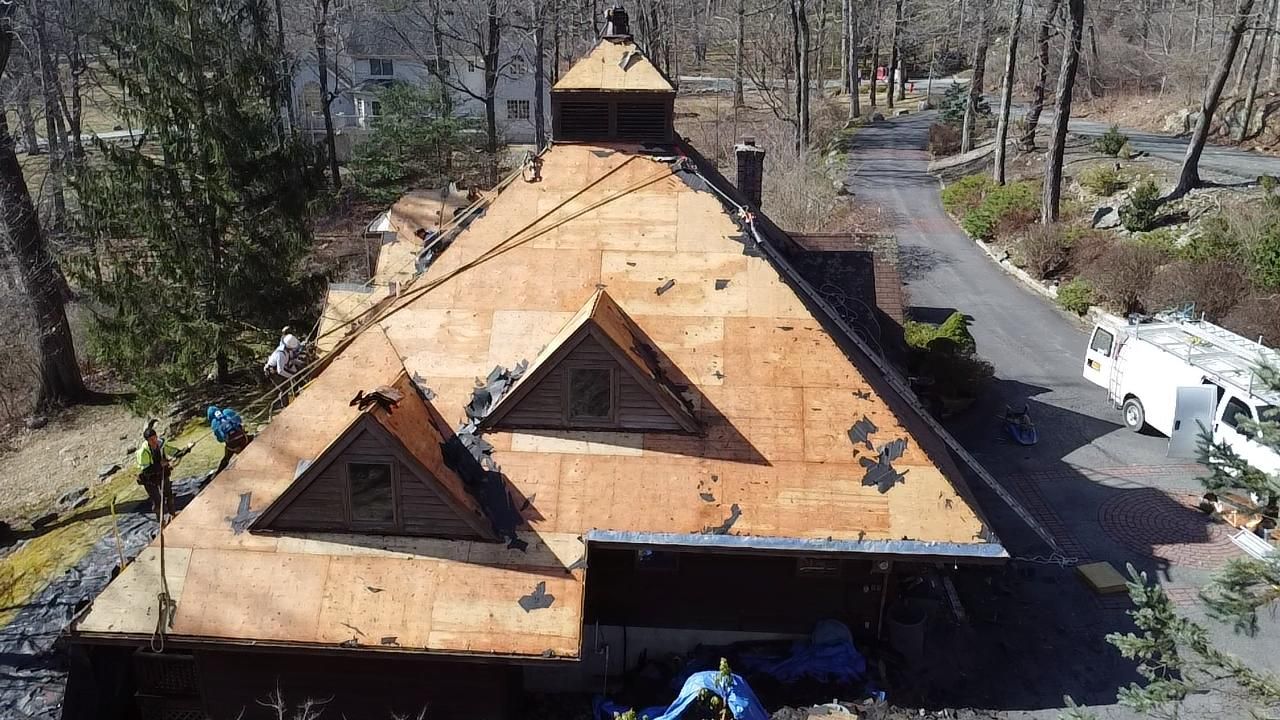 Roof of a house under construction. Brown roof with exposed underlayment. Workers are visible, surrounded by trees.