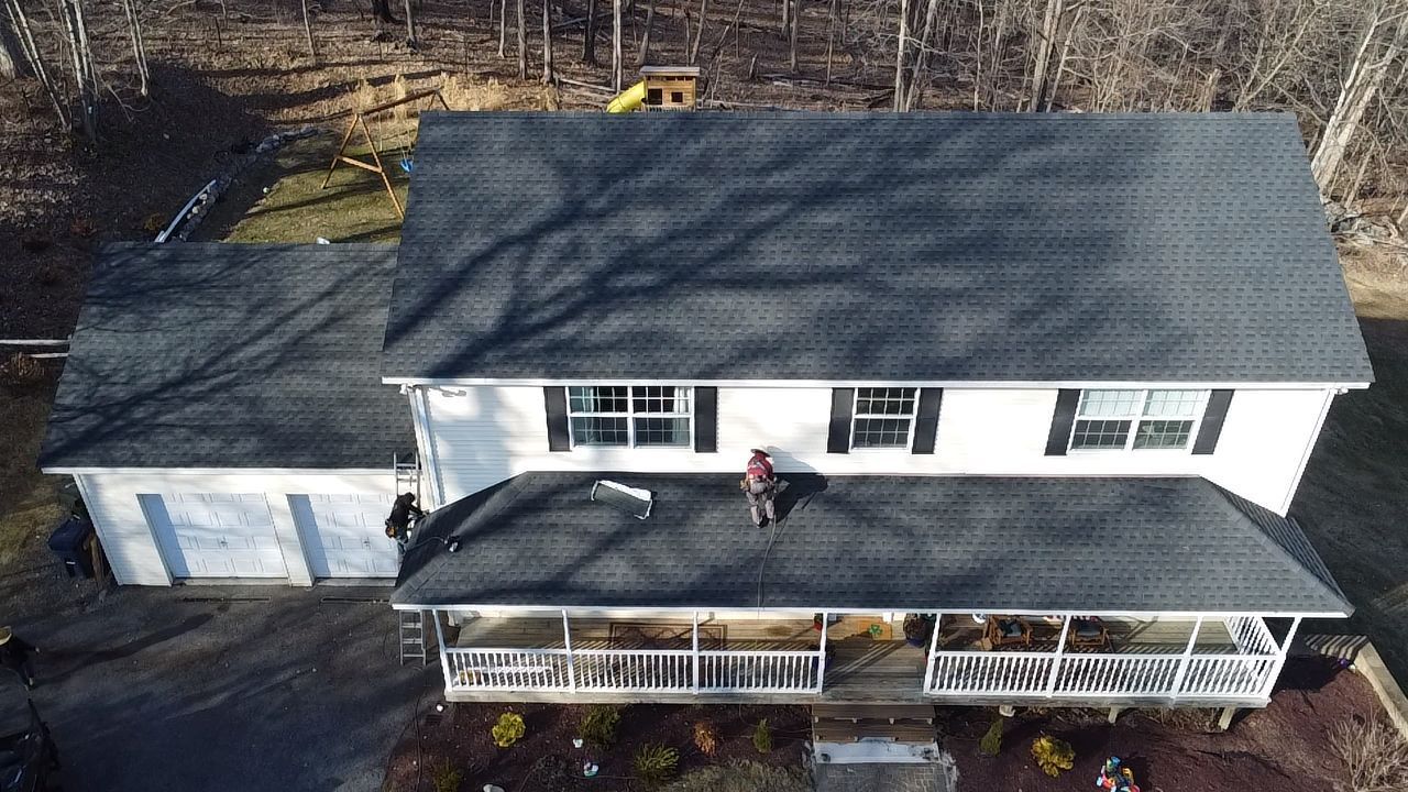 Aerial view of a white house with a dark gray roof. A person is working on the roof.