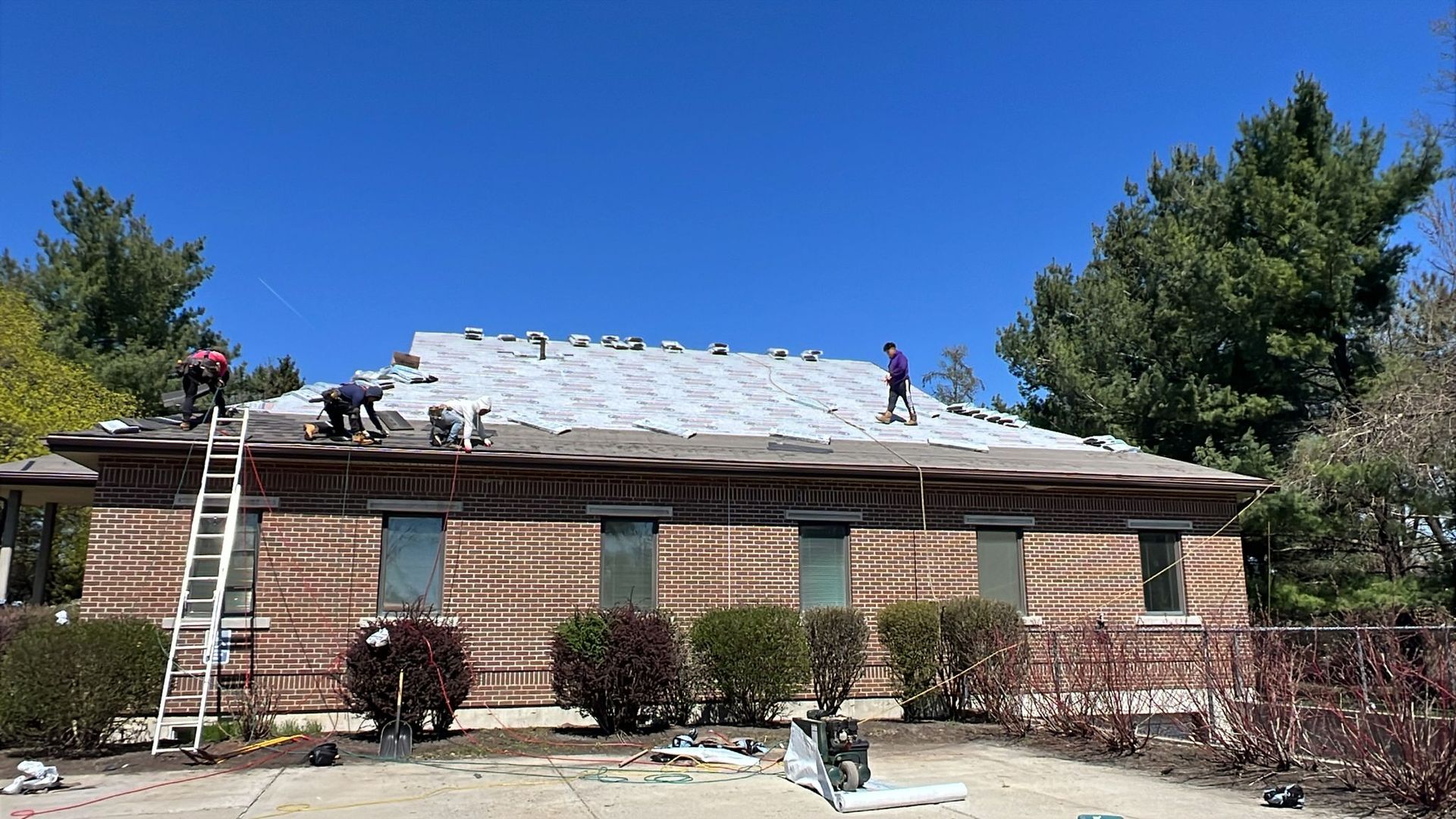 Workers on a roof, removing shingles. Brick building, ladder, blue sky.