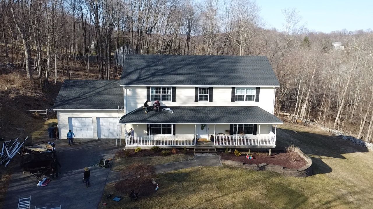 Two-story white house with black roof, covered porch, and attached garage. Men on roof, trees in background.